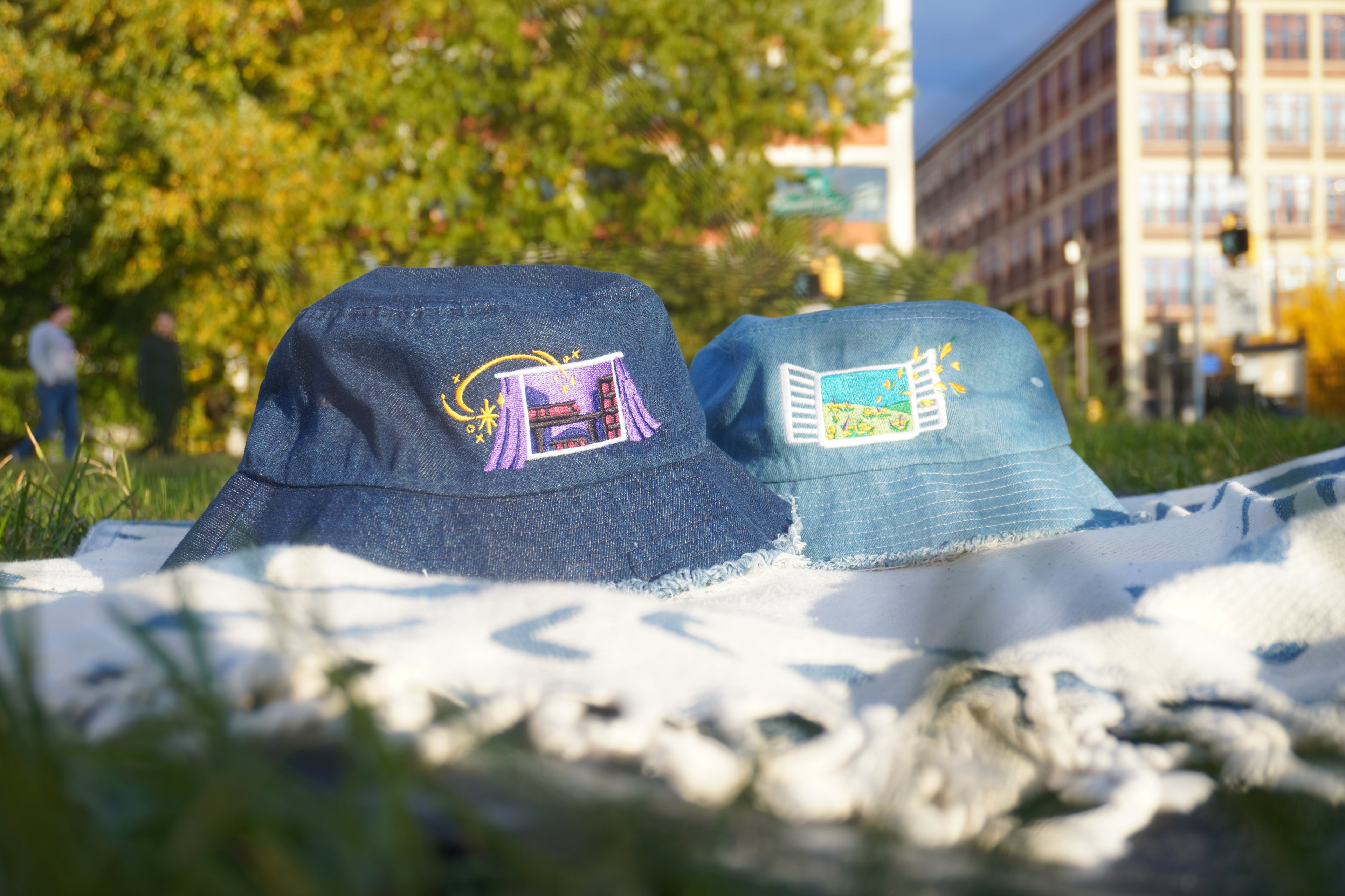 Two denim bucket hats with embroidered designs on a blanket outdoors.
