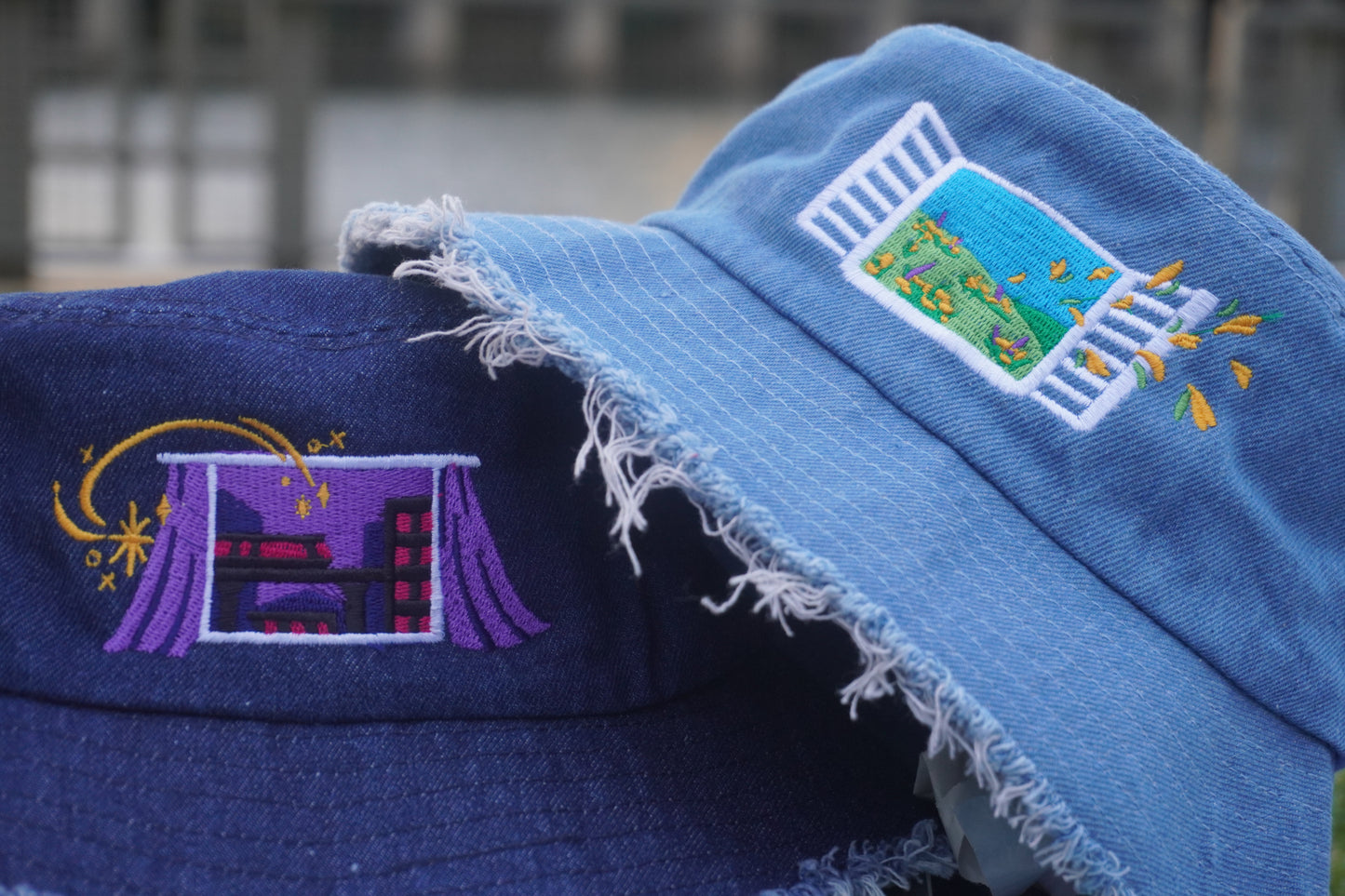 Two embroidered denim bucket hats, one light blue and one dark blue, with decorative designs against a blurred background.