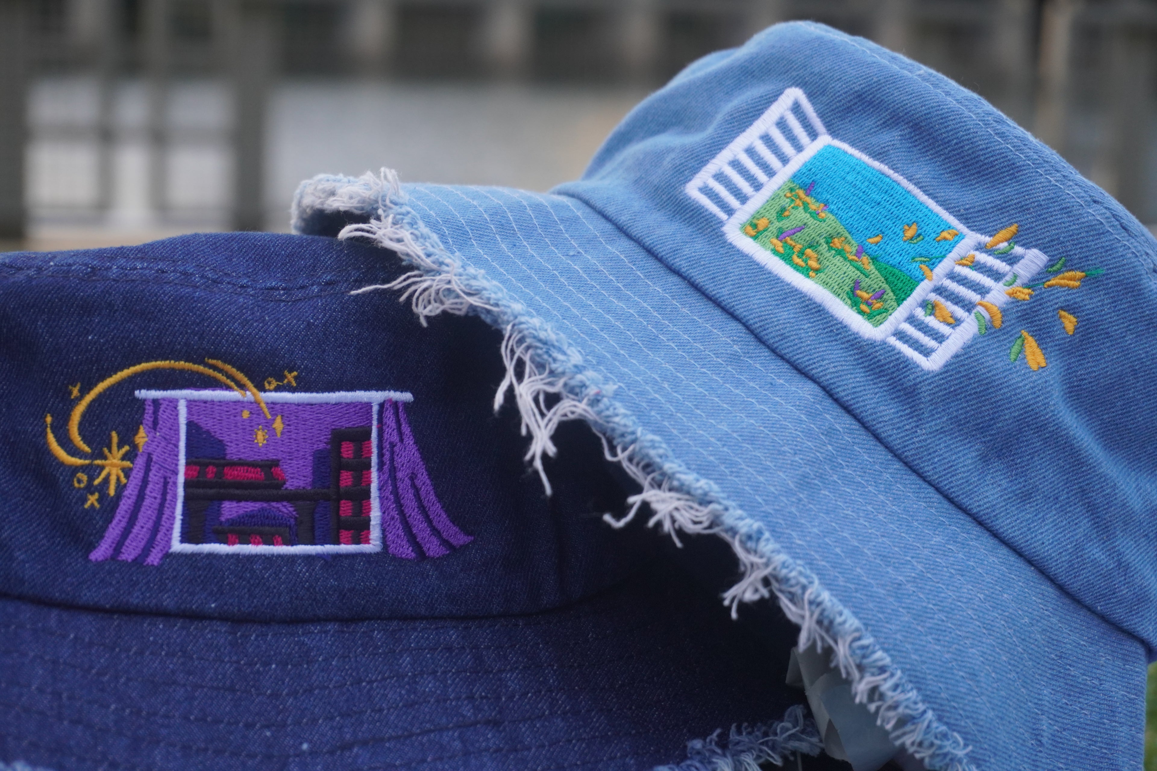 Two embroidered denim bucket hats, one light blue and one dark blue, with decorative designs against a blurred background.