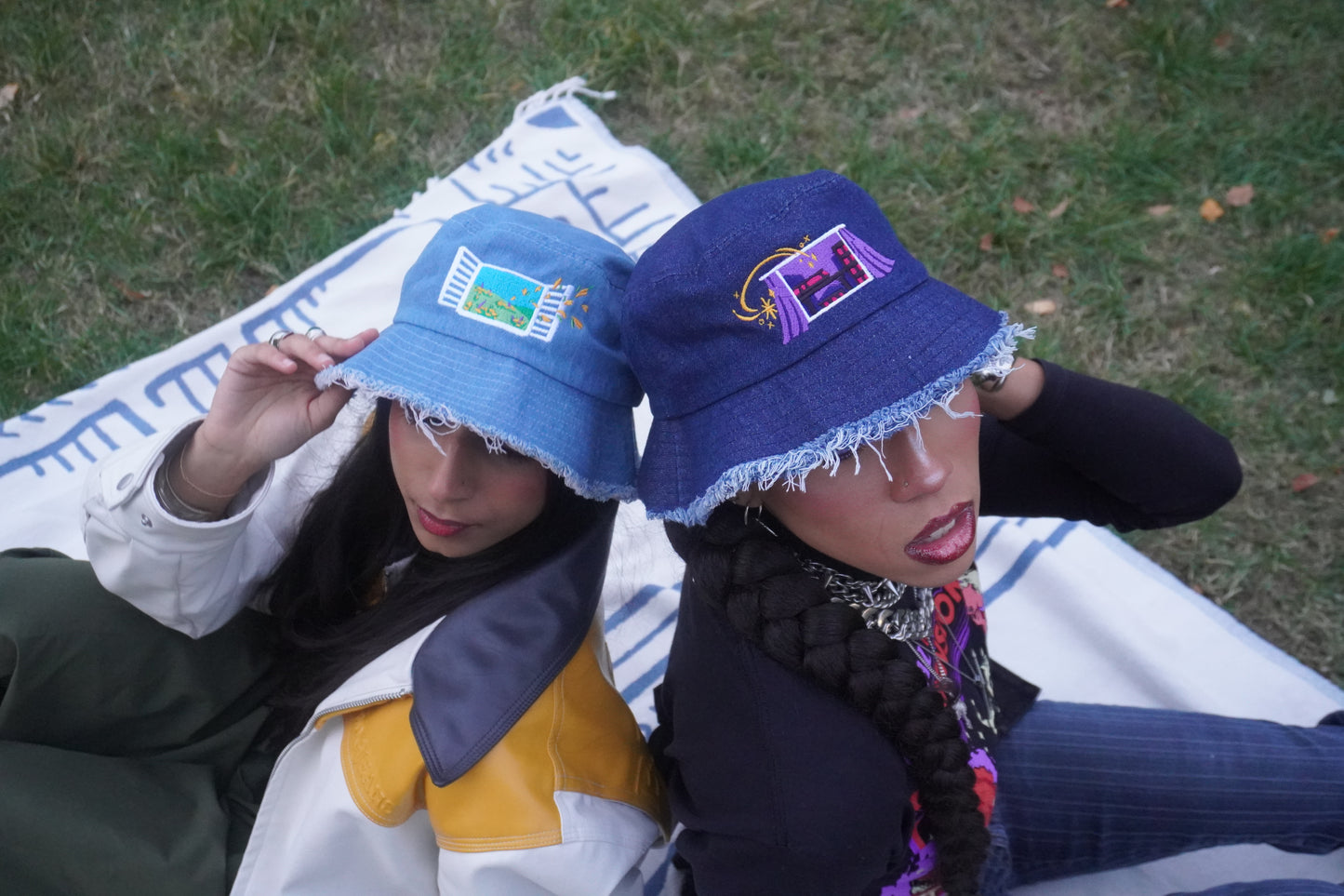 Two people wearing blue bucket hats with colorful embroidered designs, sitting on a picnic blanket in grassy area.
