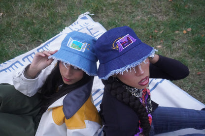 Two people wearing blue bucket hats with colorful embroidered designs, sitting on a picnic blanket in grassy area.