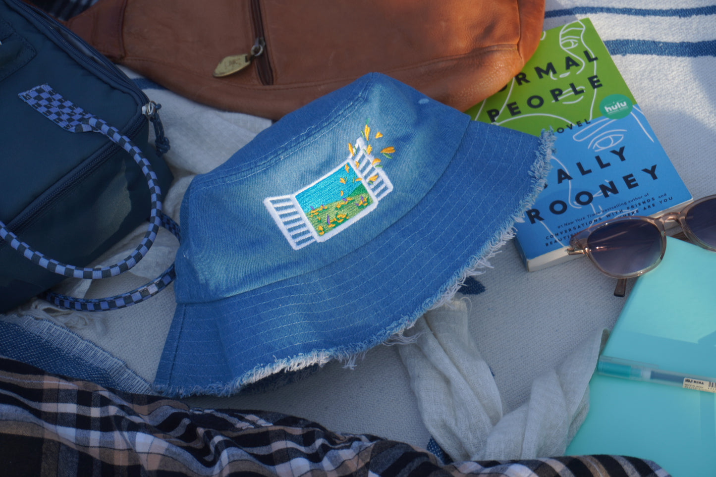 Blue bucket hat with a logo on a picnic blanket with books and sunglasses