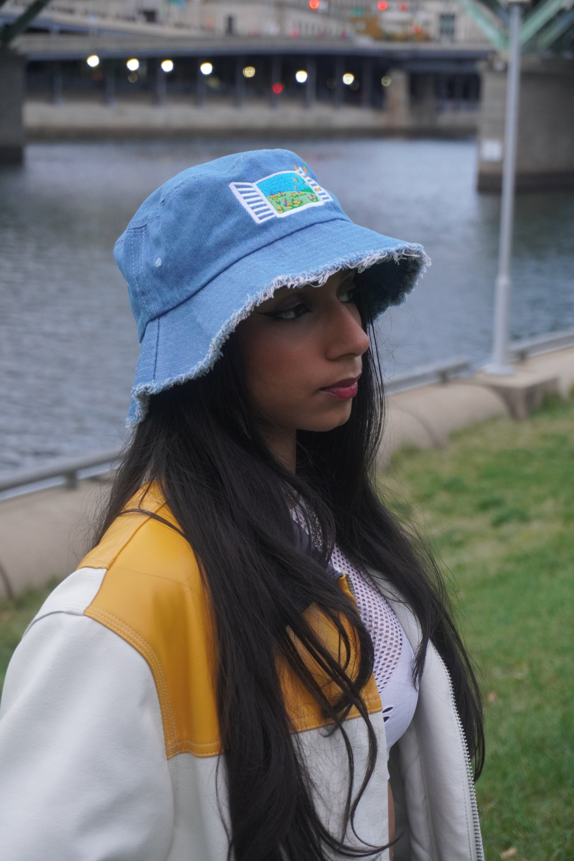 Woman wearing a blue cap with an embroidered design, white jacket with yellow accents, and long dark hair. Background is blurred.