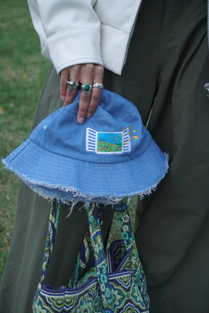 Person holding a blue bucket hat with embroidered design, wearing a white jacket, green pants, and silver and green rings, on a grassy background.