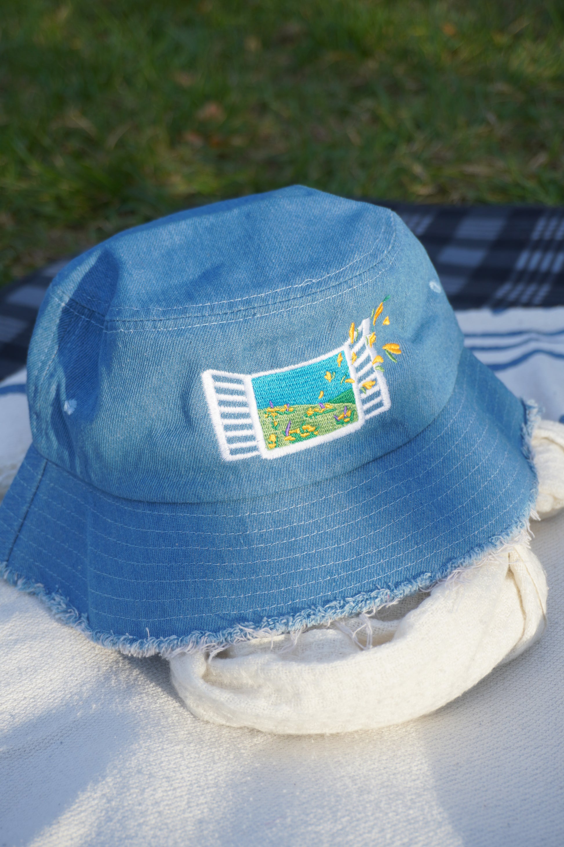 Blue denim bucket hat with embroidered design on a white picnic blanket and grassy background