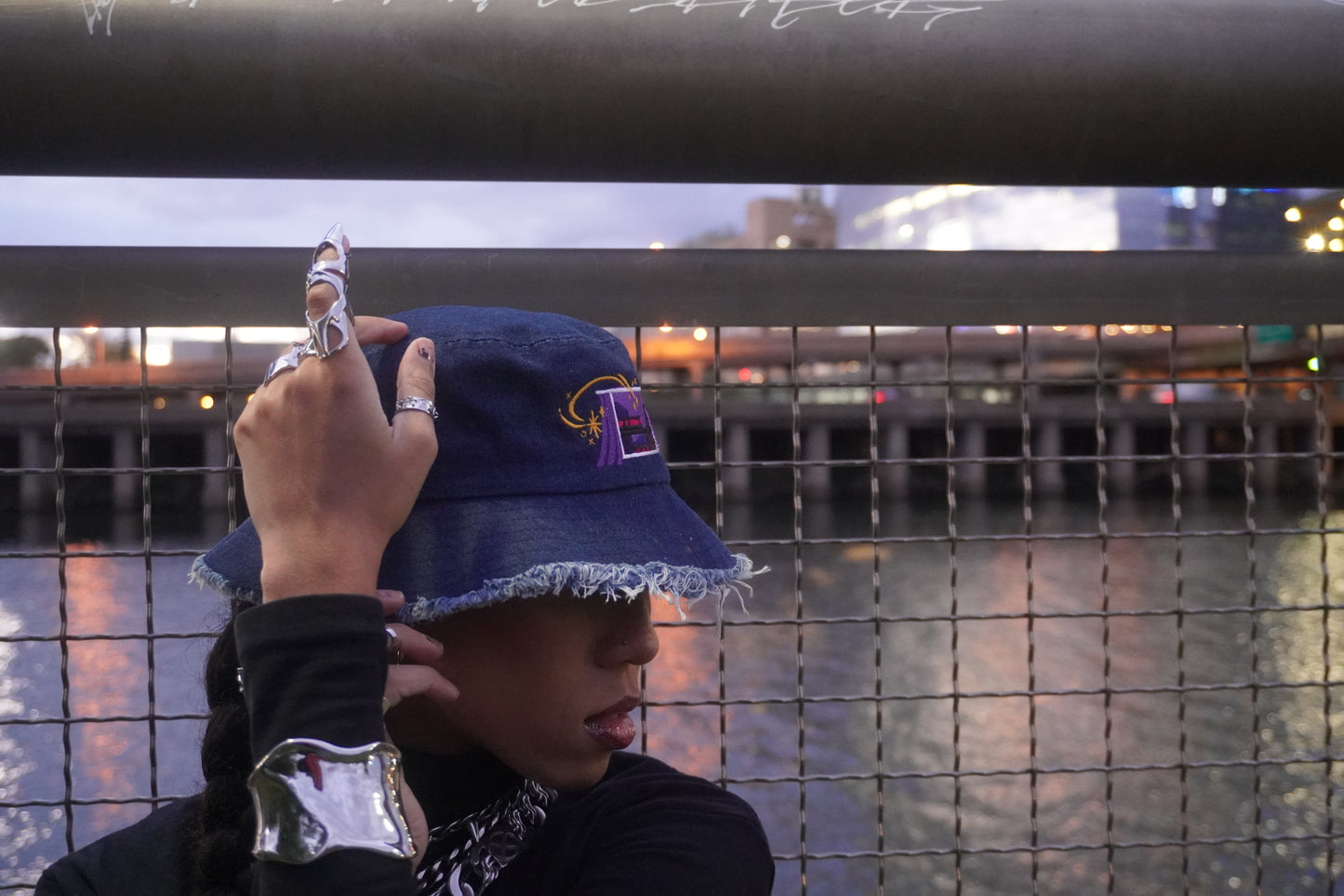 Person wearing a blue bucket hat with a logo, against a blurred background of water and lights.