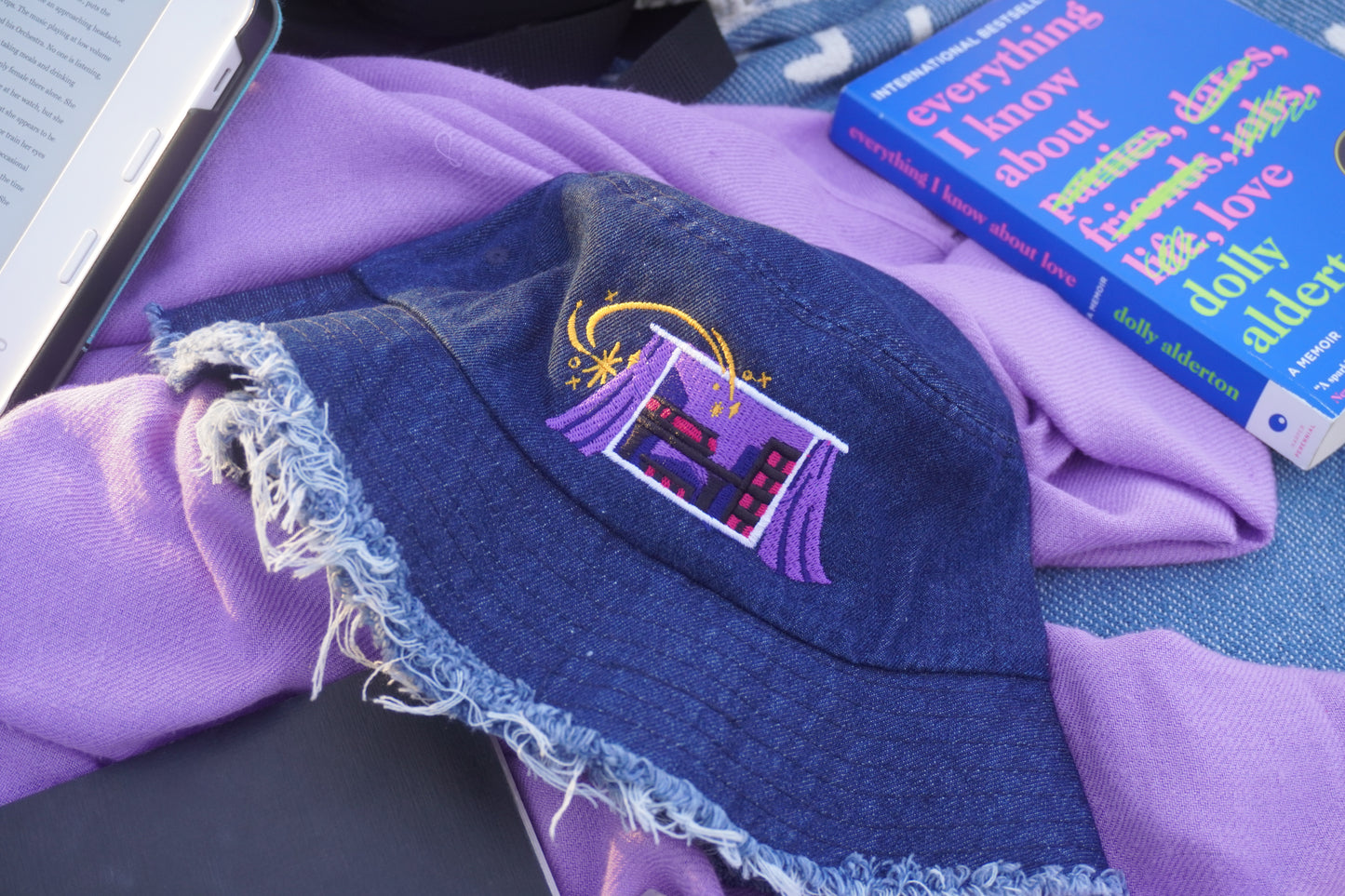 Blue denim bucket hat with embroidered design on a picnic blanket background with a purple scarf, blue book, and e-reader
