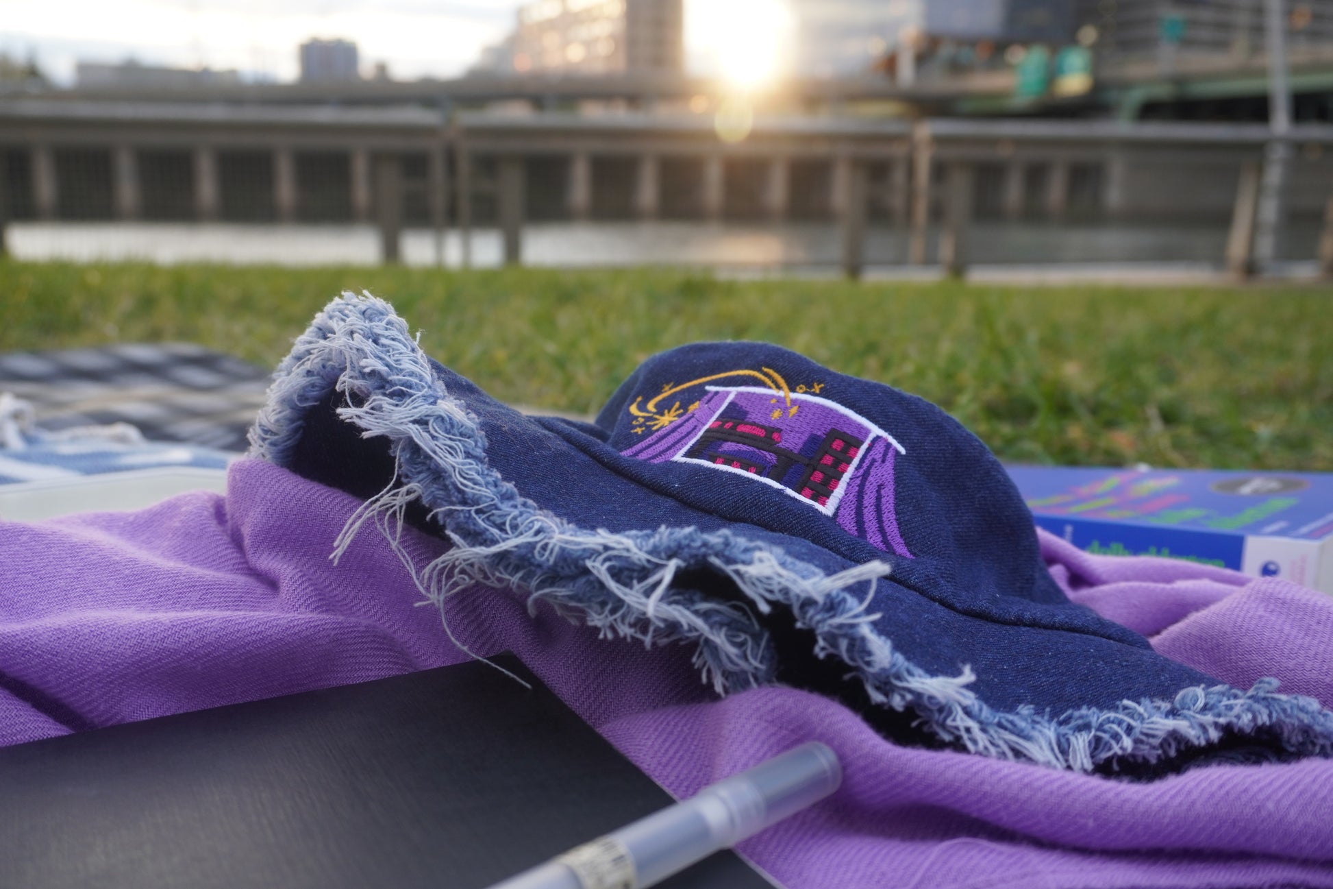 Blue bucket hat with unhemmed fringe on a purple scarf by a waterfront and grassy background.