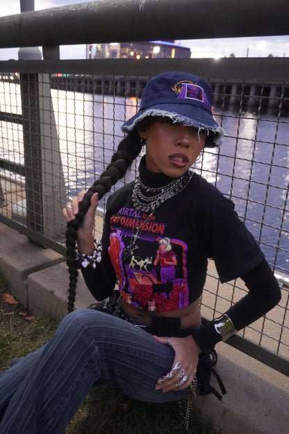 Person wearing a dark blue denim bucket hat, wearing a graphic black shirt, sitting against a railing near a river.