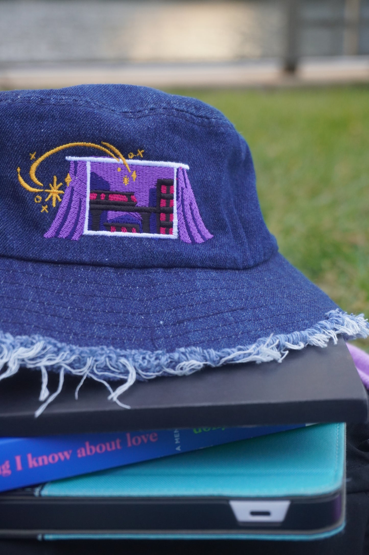 Blue denim bucket hat with embroidered design stacked on top of books and notebooks against a blurred grassy background